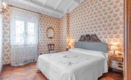 Cozy vintage bedroom featuring floral wallpaper, a decorative mirror, and a wooden chair. The room includes a plush bed with a white coverlet and elegant lighting, creating a warm and inviting atmosphere.