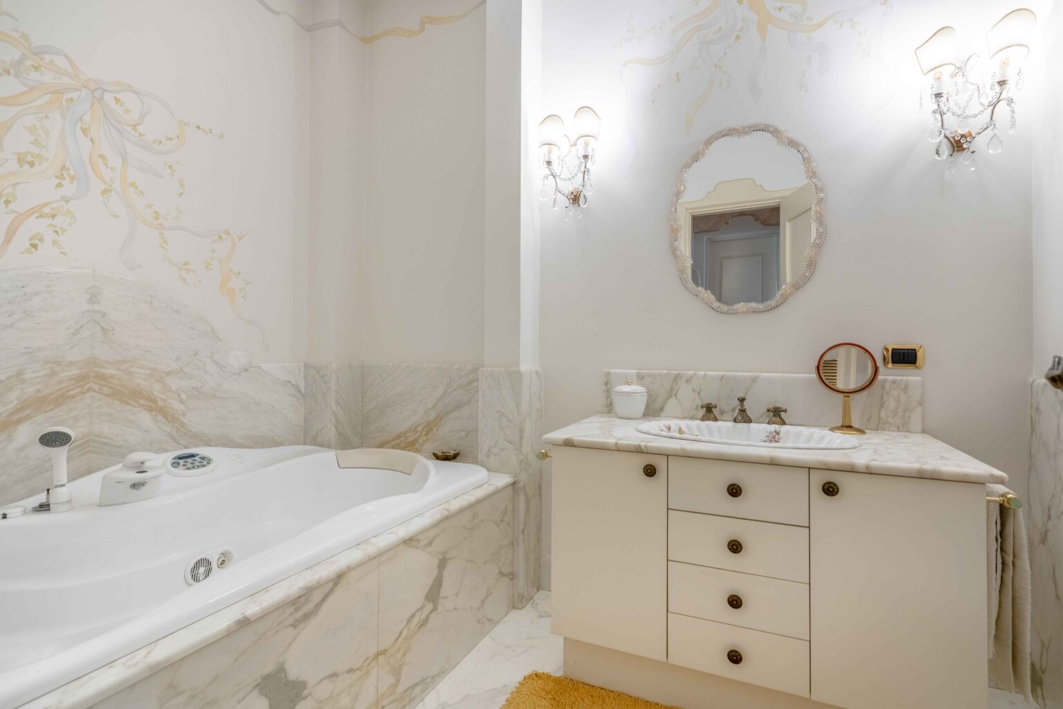 Luxurious bathroom featuring a white jacuzzi tub, elegant marble walls, and a stylish vanity with a round mirror and gold fixtures. Soft lighting enhances the serene atmosphere, complemented by decorative wall art and a plush rug. | Only on SopranoVillas