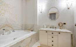 Luxurious bathroom featuring a white jacuzzi tub, elegant marble walls, and a stylish vanity with a round mirror and gold fixtures. Soft lighting enhances the serene atmosphere, complemented by decorative wall art and a plush rug.