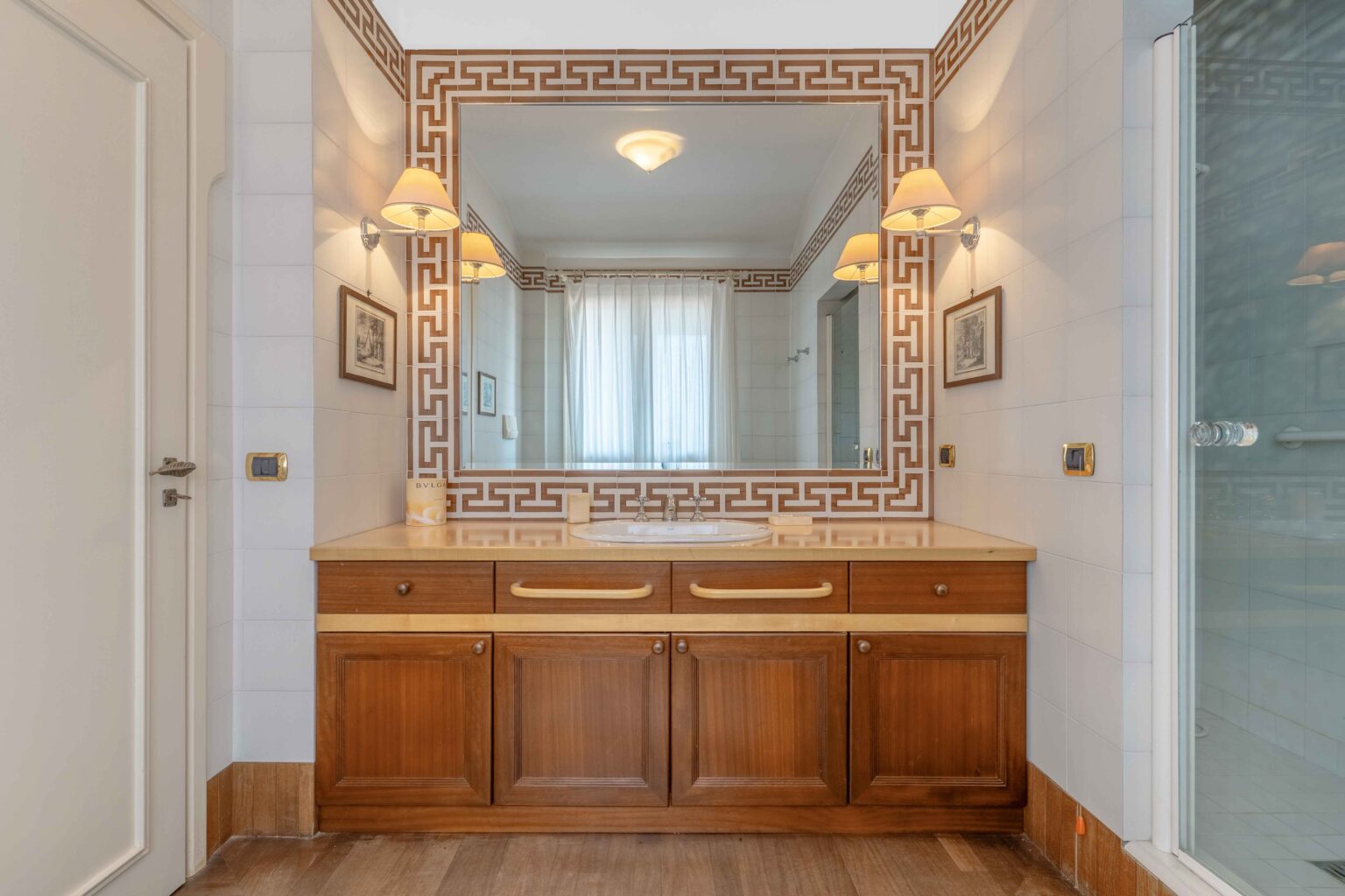 Modern bathroom interior featuring a wooden vanity with a double sink, elegant wall-mounted light fixtures, and a large mirror framed with a decorative border. The space is brightened by natural light filtering through sheer curtains, complemented by tiled walls and a glass shower enclosure. | Only on SopranoVillas
