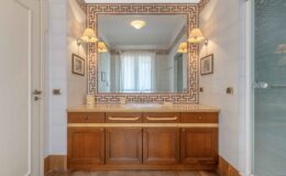 Modern bathroom interior featuring a wooden vanity with a double sink, elegant wall-mounted light fixtures, and a large mirror framed with a decorative border. The space is brightened by natural light filtering through sheer curtains, complemented by tiled walls and a glass shower enclosure.