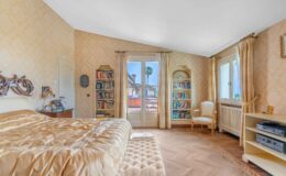 Cozy bedroom featuring a luxurious golden bedspread, elegant decor, and a bookshelf filled with books. Natural light streams through large windows, revealing a view of palm trees outside. The room includes a stylish armchair and a media console, creating a warm and inviting atmosphere.
