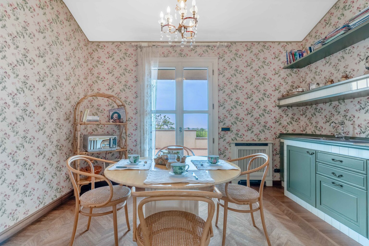 Cozy dining area featuring a round table set with floral tea cups, surrounded by bentwood chairs. The room has vintage floral wallpaper and a wooden shelf displaying books and decor, with a large window providing natural light and a view of the outdoors. | Only on SopranoVillas