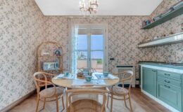 Cozy dining area featuring a round table set with floral tea cups, surrounded by bentwood chairs. The room has vintage floral wallpaper and a wooden shelf displaying books and decor, with a large window providing natural light and a view of the outdoors.