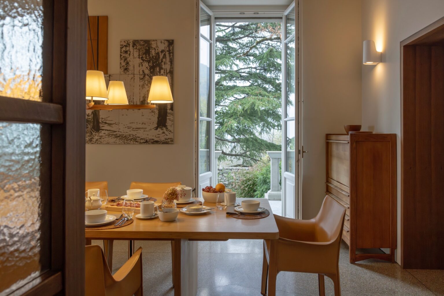Elegant dining area in a luxury villa for rent in Italy, featuring a wooden table set for breakfast, stylish chairs, and large windows offering a scenic view of the outdoors. | Only on SopranoVillas