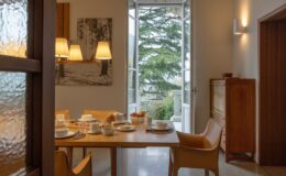 Elegant dining area in a luxury villa for rent in Italy, featuring a wooden table set for breakfast, stylish chairs, and large windows offering a scenic view of the outdoors.
