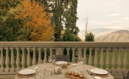Luxury villa dining terrace in Italy, featuring a beautifully set table with elegant dinnerware, surrounded by lush greenery and autumn foliage, offering stunning mountain views. Perfect for vacation rentals.