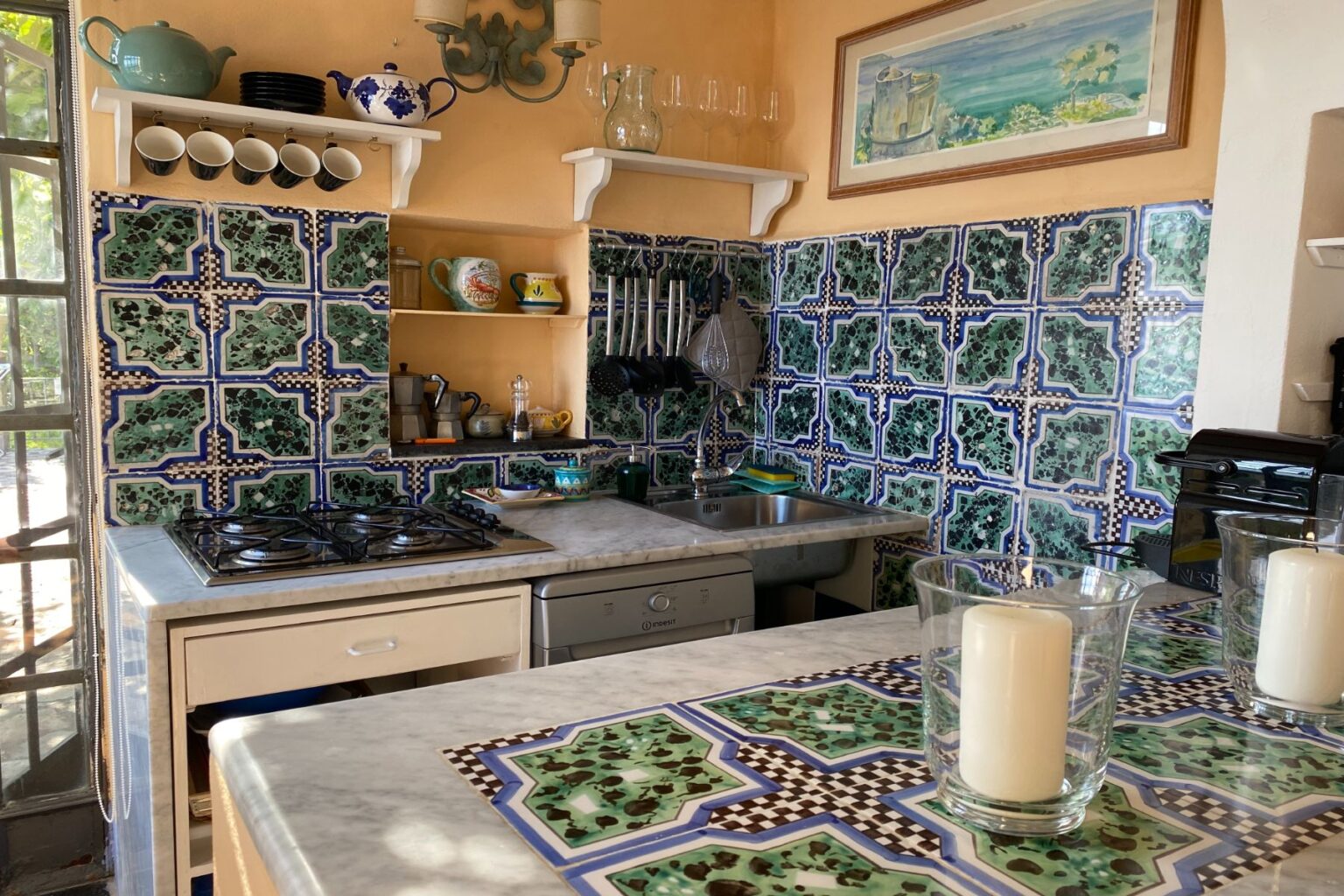 Bright and colorful kitchen featuring intricate green and blue tile backsplash, modern appliances, and a marble countertop with decorative candles. The space is well-lit and showcases a cozy, inviting atmosphere. | Only on SopranoVillas
