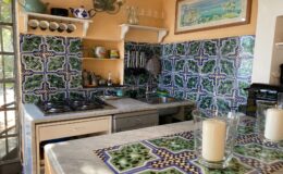 Bright and colorful kitchen featuring intricate green and blue tile backsplash, modern appliances, and a marble countertop with decorative candles. The space is well-lit and showcases a cozy, inviting atmosphere.