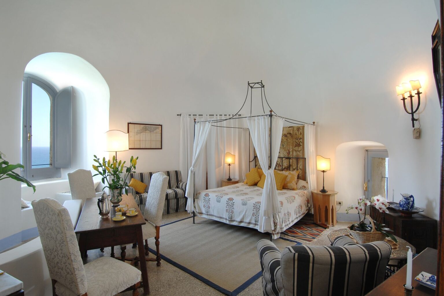 Elegant bedroom with a four-poster bed, featuring soft linens and decorative pillows. The room includes a cozy seating area, a wooden table, and large windows allowing natural light, creating a serene and inviting atmosphere. | Only on SopranoVillas