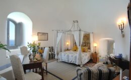 Elegant bedroom with a four-poster bed, featuring soft linens and decorative pillows. The room includes a cozy seating area, a wooden table, and large windows allowing natural light, creating a serene and inviting atmosphere.