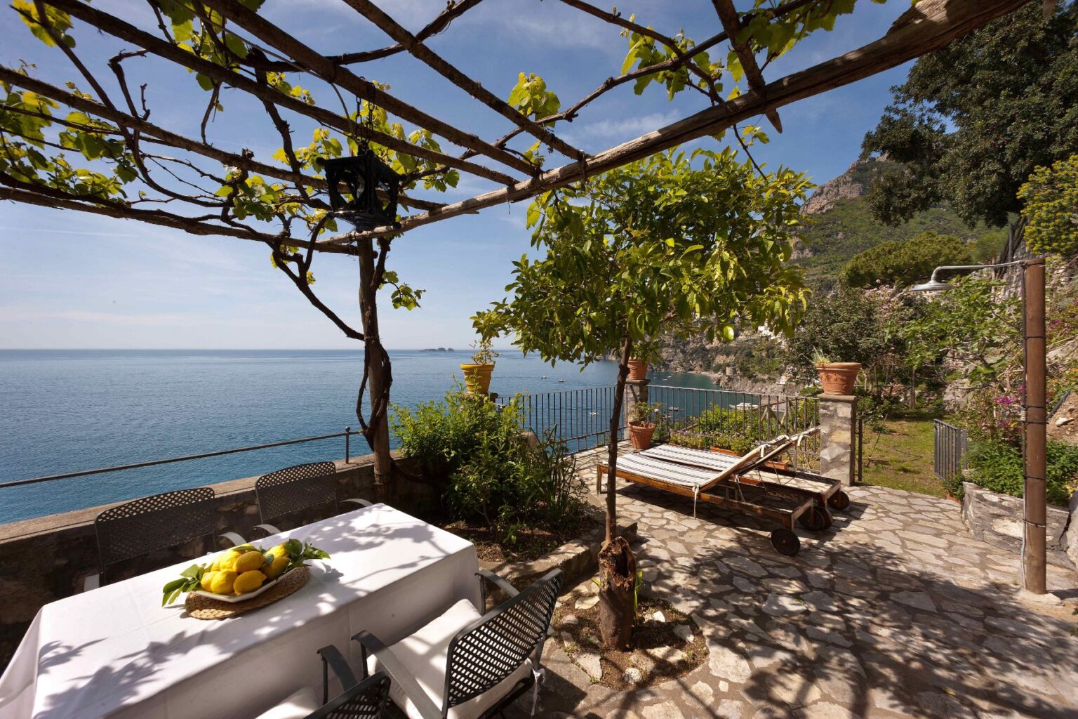 Scenic outdoor patio overlooking the ocean, featuring a dining table with fresh fruit, a sun lounger, and lush greenery under a pergola. Perfect for relaxation and enjoying coastal views. | Only on SopranoVillas