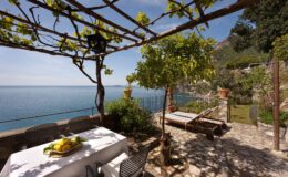 Scenic outdoor patio overlooking the ocean, featuring a dining table with fresh fruit, a sun lounger, and lush greenery under a pergola. Perfect for relaxation and enjoying coastal views.
