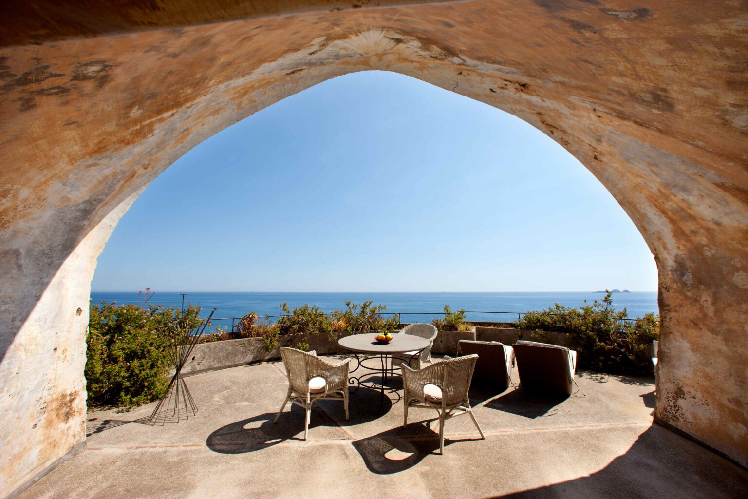Cozy outdoor seating area with a table and chairs, framed by an archway, overlooking a serene ocean view under clear blue skies. Ideal for relaxation and enjoyment of coastal scenery. | Only on SopranoVillas