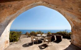 Cozy outdoor seating area with a table and chairs, framed by an archway, overlooking a serene ocean view under clear blue skies. Ideal for relaxation and enjoyment of coastal scenery.