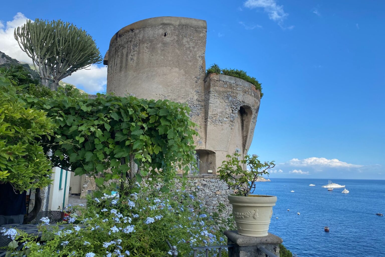 Historic coastal tower surrounded by lush greenery and blooming flowers, overlooking a serene blue sea under a clear sky. | Only on SopranoVillas