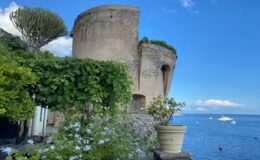 Historic coastal tower surrounded by lush greenery and blooming flowers, overlooking a serene blue sea under a clear sky.