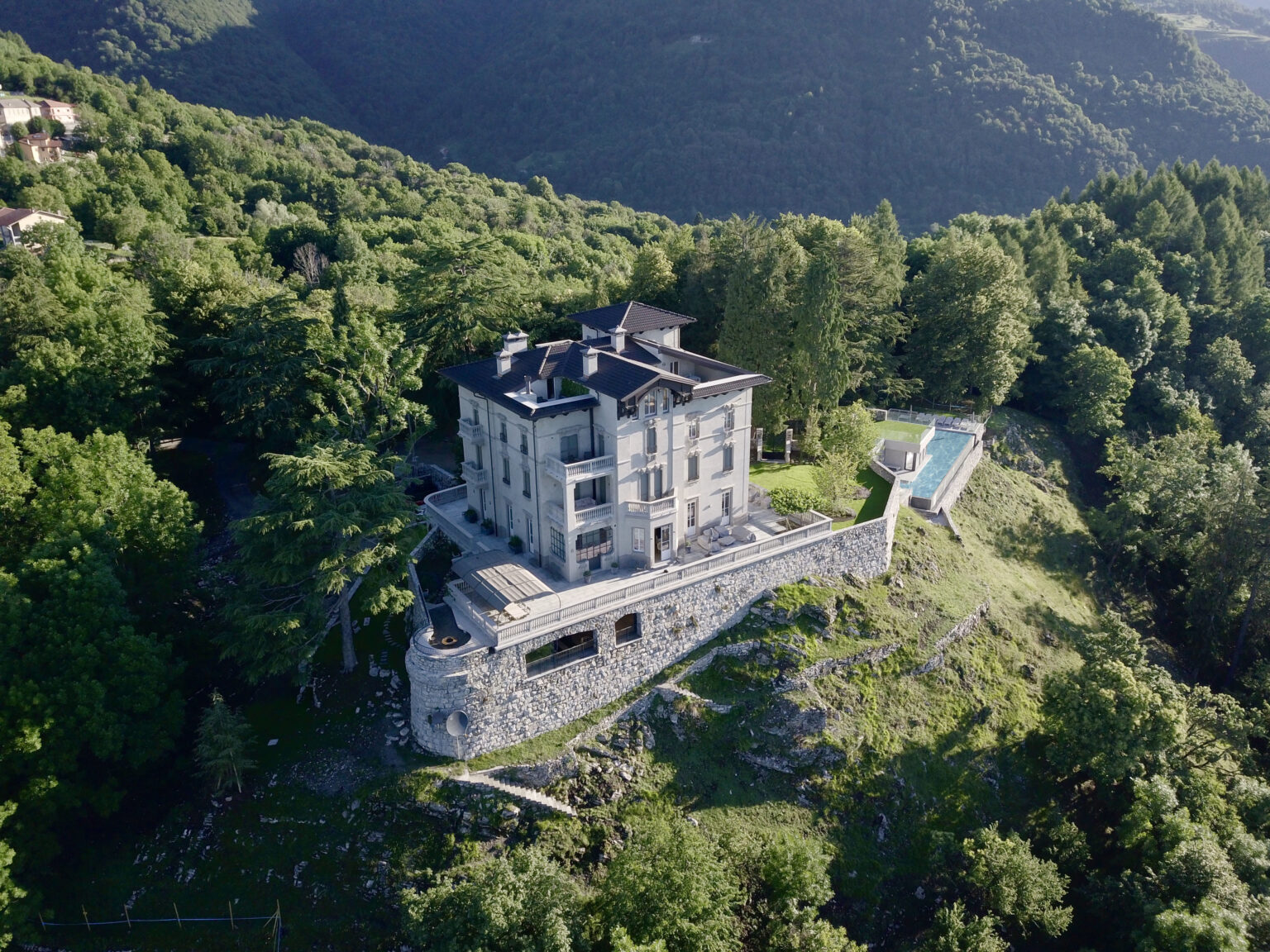 Aerial view of a luxurious modern villa surrounded by lush greenery and mountains, featuring a swimming pool and stone terrace, ideal for serene getaways. | Only on SopranoVillas