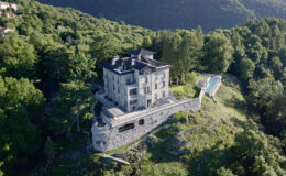 Aerial view of a luxurious modern villa surrounded by lush greenery and mountains, featuring a swimming pool and stone terrace, ideal for serene getaways.