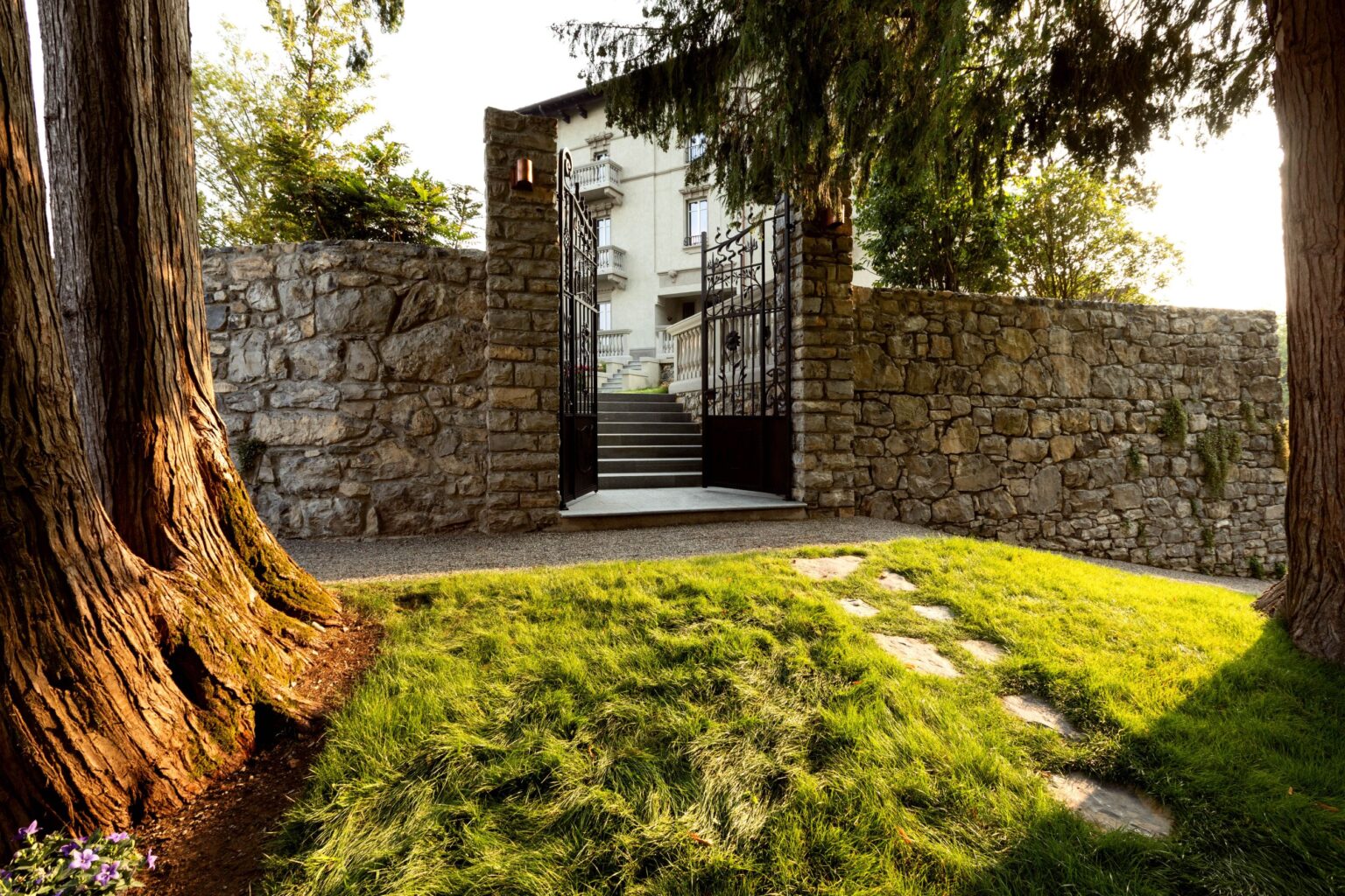 Stone wall entrance with a gated pathway leading to a modern building, surrounded by lush green grass and tall trees, creating a serene outdoor space. | Only on SopranoVillas
