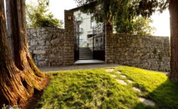 Stone wall entrance with a gated pathway leading to a modern building, surrounded by lush green grass and tall trees, creating a serene outdoor space.