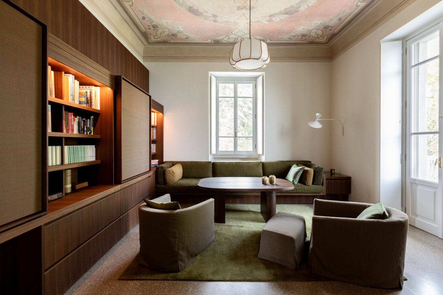 Modern living room interior featuring a cozy green sofa, stylish armchairs, and a round table, complemented by a decorative ceiling and natural light from large windows. | Only on SopranoVillas