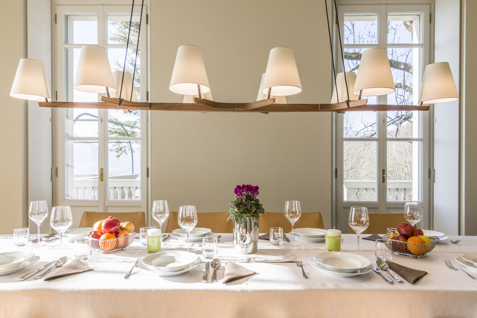 Elegant dining table set for a meal, featuring a neutral backdrop with large windows. The table is adorned with glassware, cutlery, colorful fruit arrangements, and a floral centerpiece, illuminated by stylish pendant lighting. Perfect for hosting gatherings or special occasions. | Only on SopranoVillas