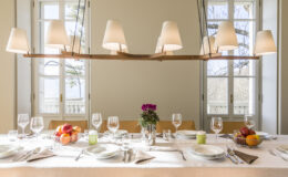 Elegant dining table set for a meal, featuring a neutral backdrop with large windows. The table is adorned with glassware, cutlery, colorful fruit arrangements, and a floral centerpiece, illuminated by stylish pendant lighting. Perfect for hosting gatherings or special occasions.