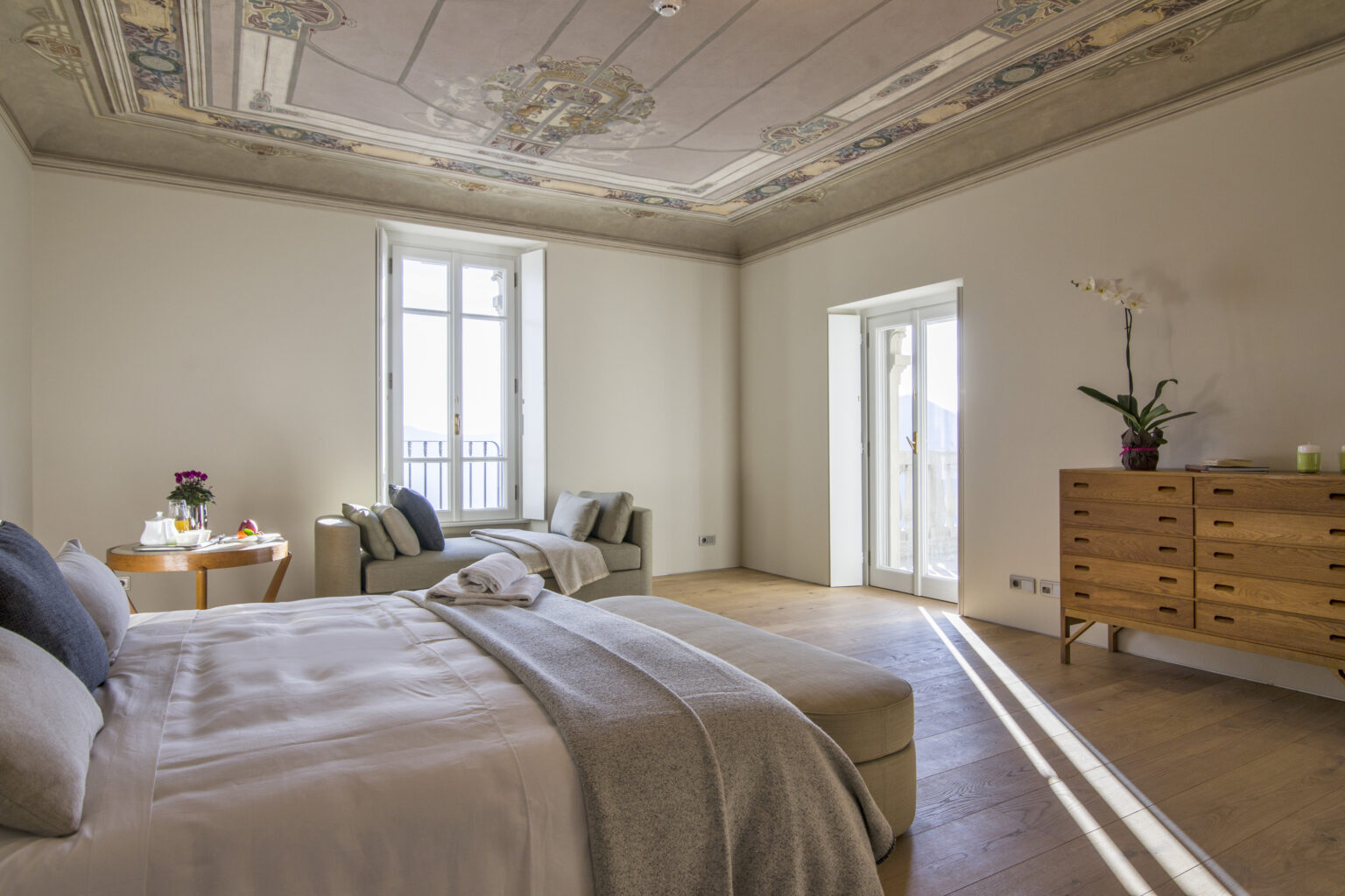 Spacious bedroom featuring a stylish bed with neutral bedding, modern wooden furniture, a cozy seating area, and large windows allowing natural light, complemented by elegant ceiling details and indoor plants. | Only on SopranoVillas
