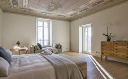 Spacious bedroom featuring a stylish bed with neutral bedding, modern wooden furniture, a cozy seating area, and large windows allowing natural light, complemented by elegant ceiling details and indoor plants.