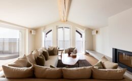 Spacious modern living room featuring a large L-shaped beige sofa, wooden coffee table, and large windows providing natural light, creating a warm and inviting atmosphere.