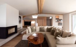 Modern living room featuring a spacious beige sectional sofa, a wooden coffee table, and a cozy fireplace. The room is bright with natural light and has wooden beams accentuating the ceiling, creating a warm and inviting atmosphere.