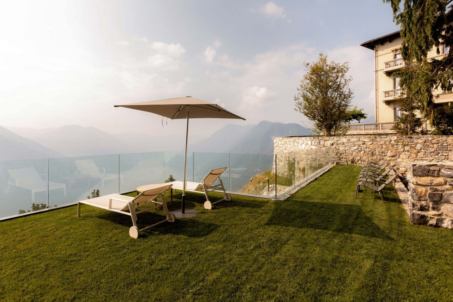 Modern outdoor terrace with sun loungers and a large umbrella, overlooking a scenic mountain landscape. The area features well-maintained green grass and a stone wall, creating a relaxing atmosphere for leisure and enjoyment. | Only on SopranoVillas