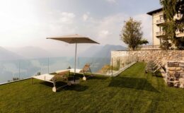 Modern outdoor terrace with sun loungers and a large umbrella, overlooking a scenic mountain landscape. The area features well-maintained green grass and a stone wall, creating a relaxing atmosphere for leisure and enjoyment.