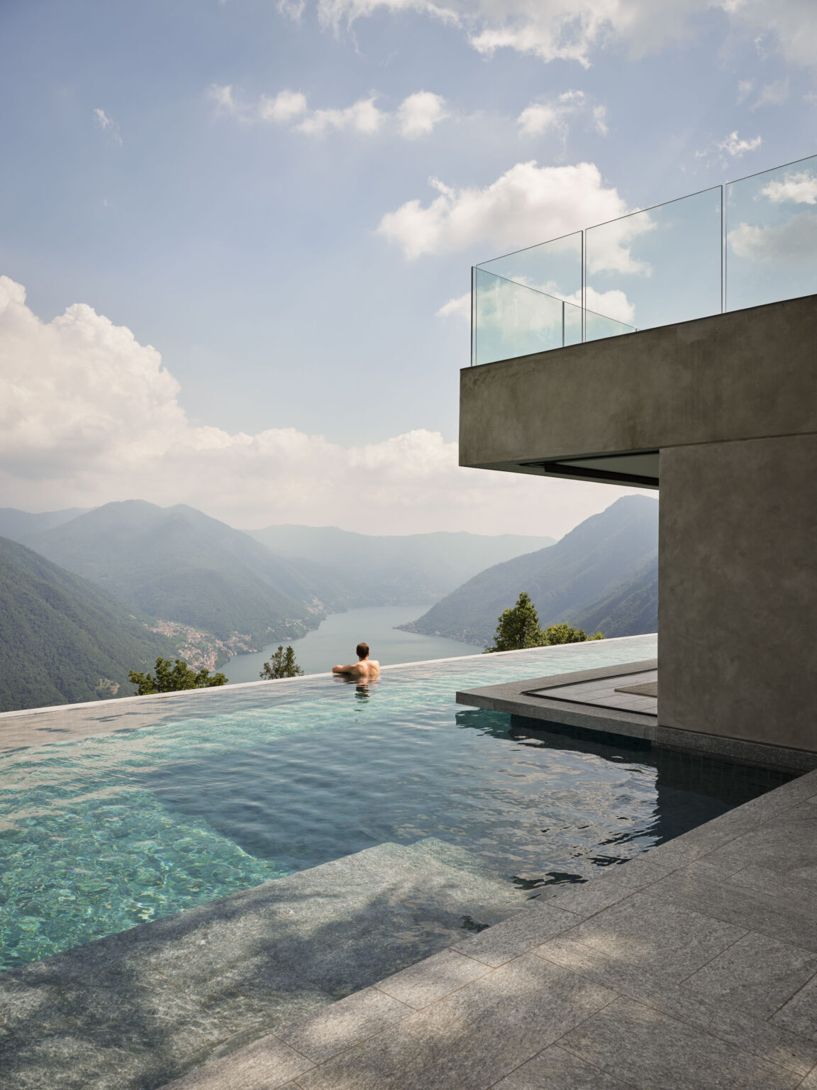 Person relaxing in an infinity pool overlooking a scenic mountain landscape and a lake, with modern architectural elements in the foreground. | Only on SopranoVillas