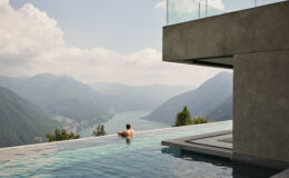 Person relaxing in an infinity pool overlooking a scenic mountain landscape and a lake, with modern architectural elements in the foreground.