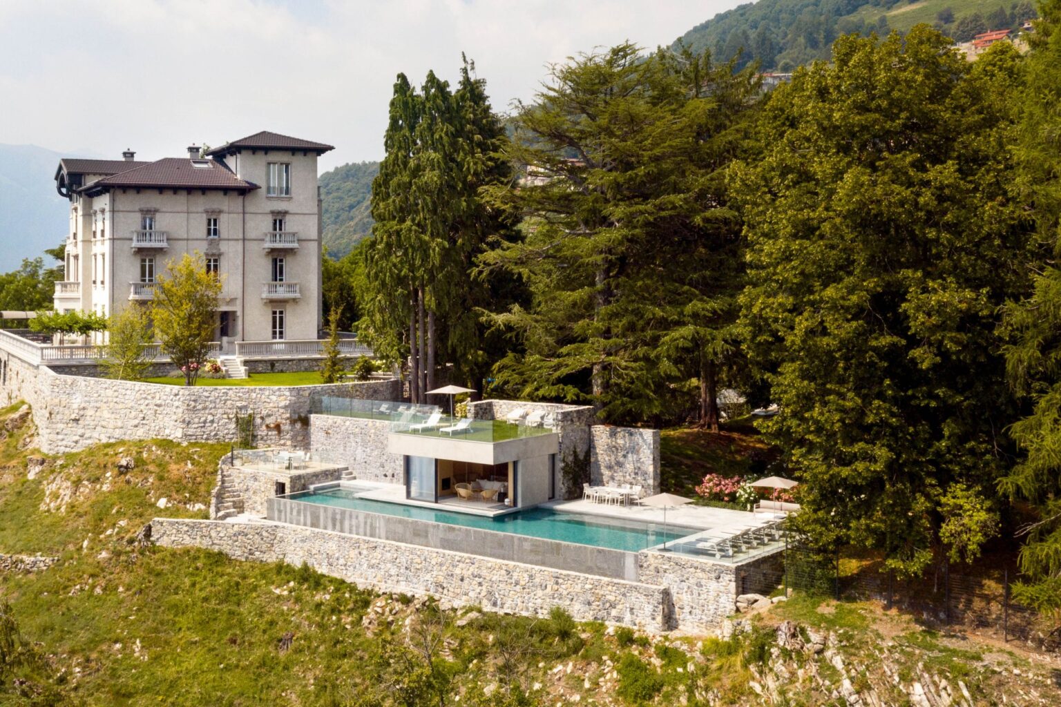 Luxury hillside villa with a modern design featuring a stone terrace, infinity pool, and lush greenery, surrounded by scenic mountains. | Only on SopranoVillas