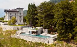 Luxury hillside villa with a modern design featuring a stone terrace, infinity pool, and lush greenery, surrounded by scenic mountains.