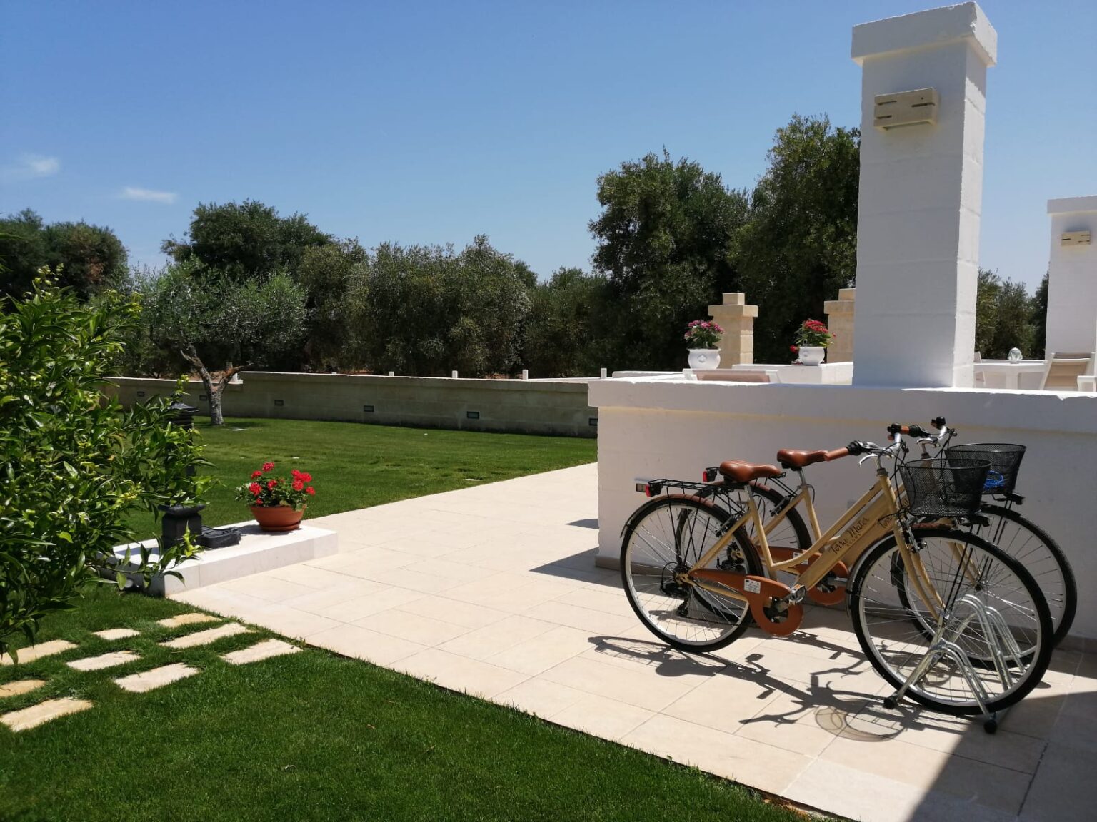 Bicycles parked on a sunlit patio surrounded by lush greenery and blooming flowers, with a clear blue sky and olive trees in the background, ideal for outdoor relaxation and exploration. | Only on SopranoVillas