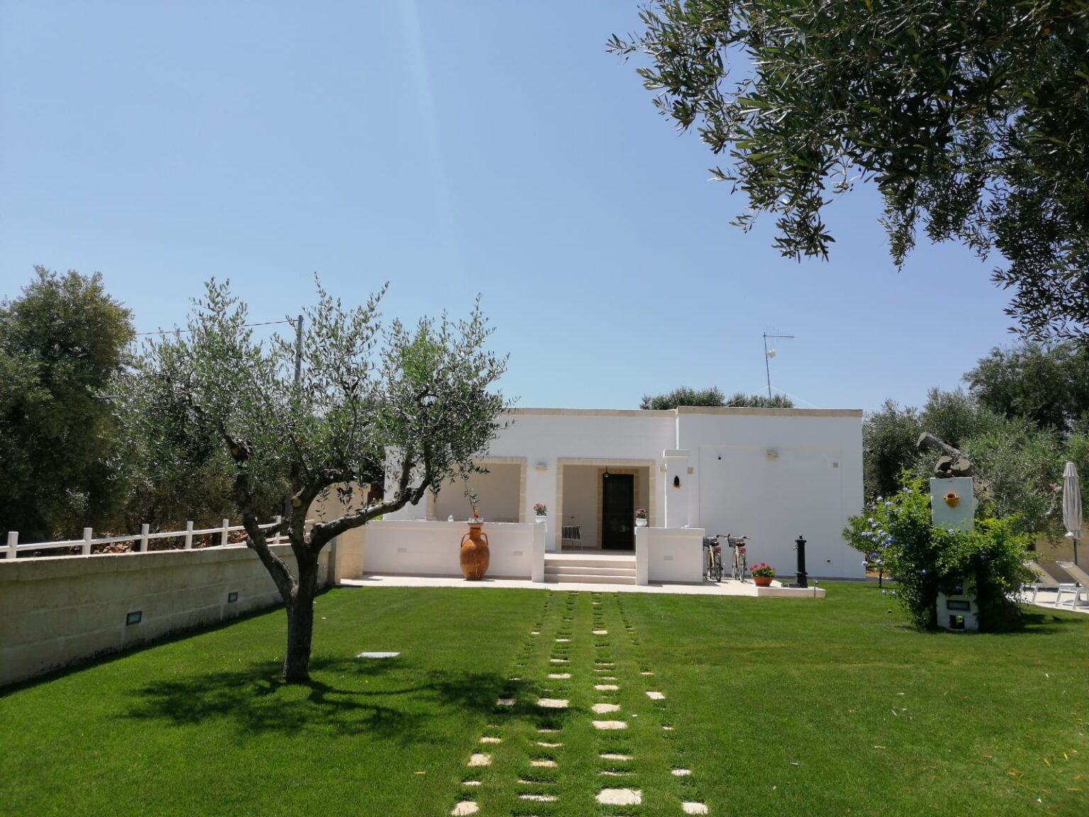 White modern villa surrounded by lush green grass and olive trees, featuring a stone pathway leading to the entrance, an outdoor seating area, and decorative elements like a large clay pot and bicycles. Clear blue sky enhances the tranquil setting. | Only on SopranoVillas