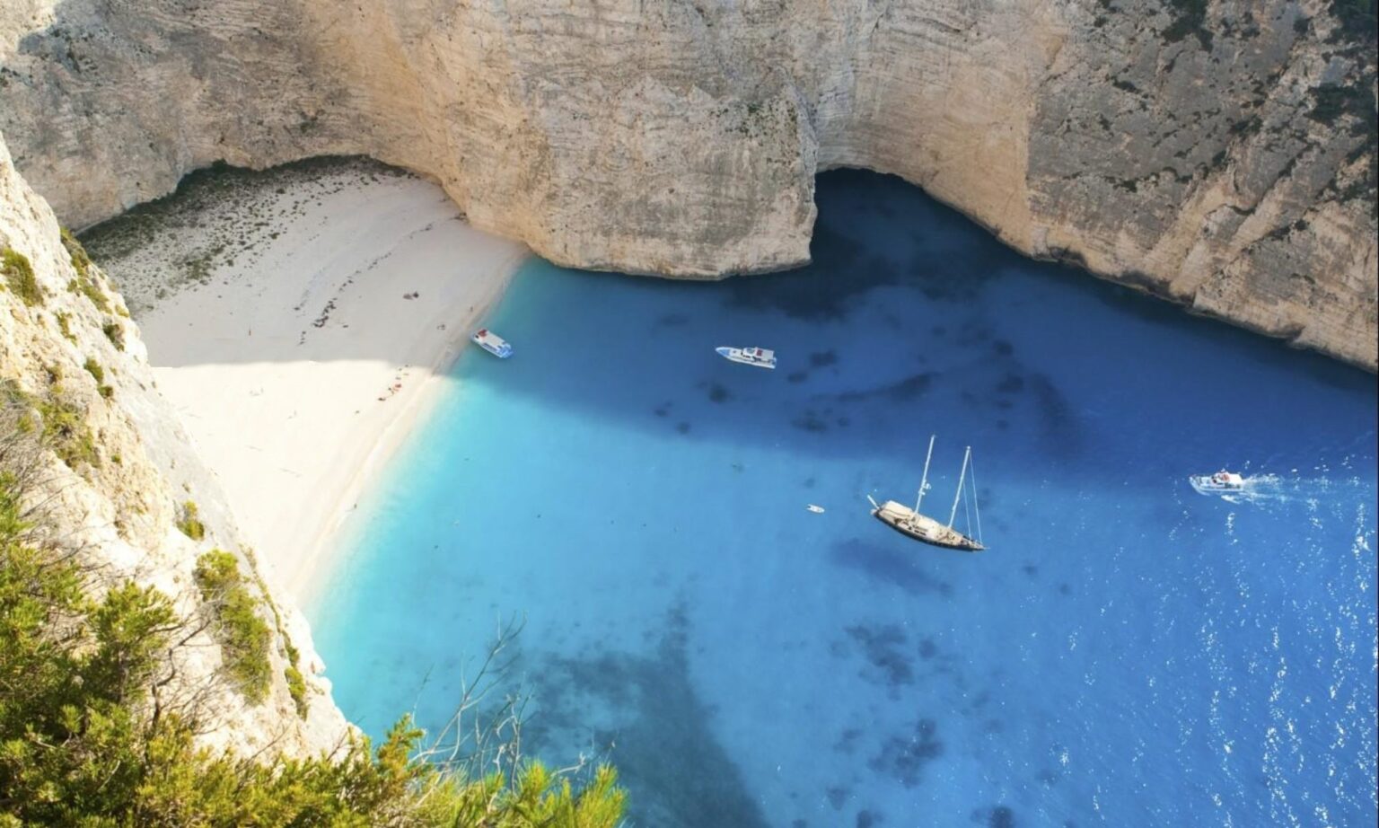 Aerial view of Navagio Beach in Zakynthos, Greece, featuring a secluded sandy cove surrounded by steep cliffs, crystal-clear turquoise waters, and several boats anchored in the bay. | Only on SopranoVillas