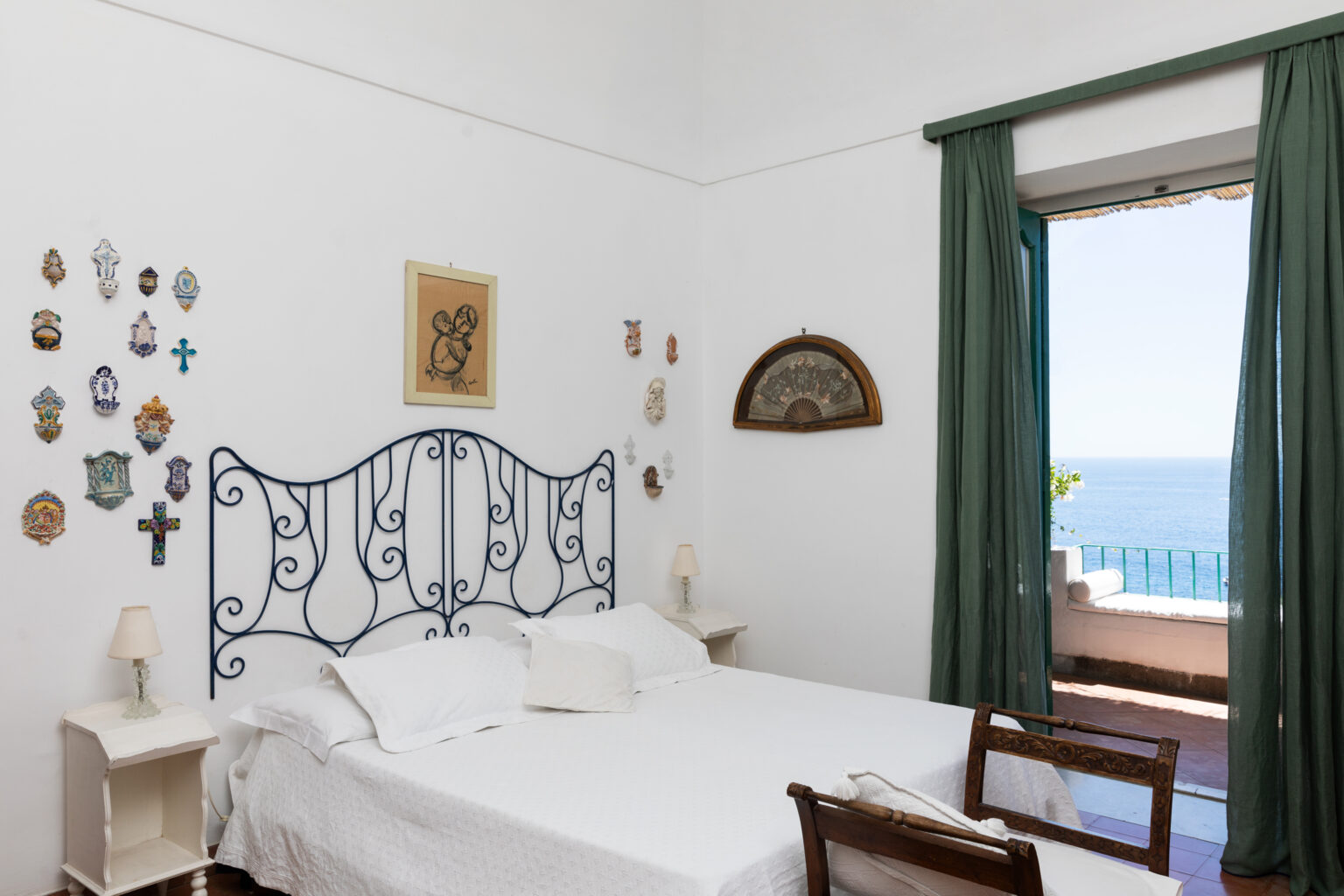 Cozy bedroom featuring a white iron bed, decorative wall art, and large windows with green curtains overlooking a scenic view of the sea. Ideal for relaxation and tranquility. | Only on SopranoVillas