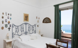 Cozy bedroom featuring a white iron bed, decorative wall art, and large windows with green curtains overlooking a scenic view of the sea. Ideal for relaxation and tranquility.