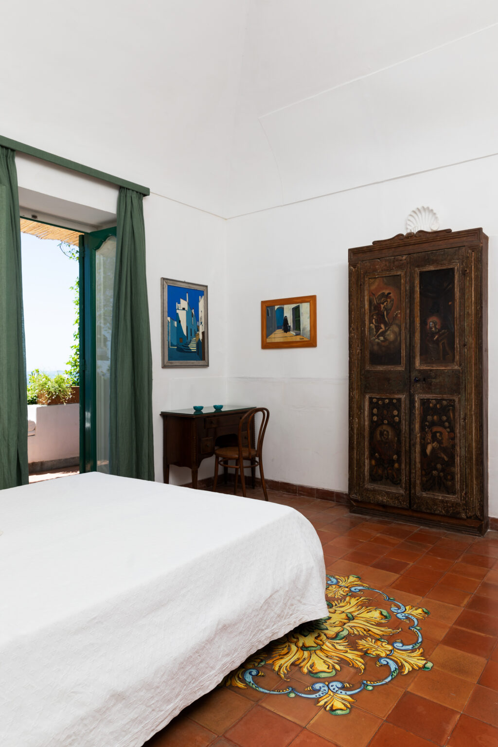 Cozy bedroom featuring a vintage wooden wardrobe, a small desk with a chair, and framed artwork on the walls. Large windows with green curtains allow natural light and a view of the outdoors. | Only on SopranoVillas