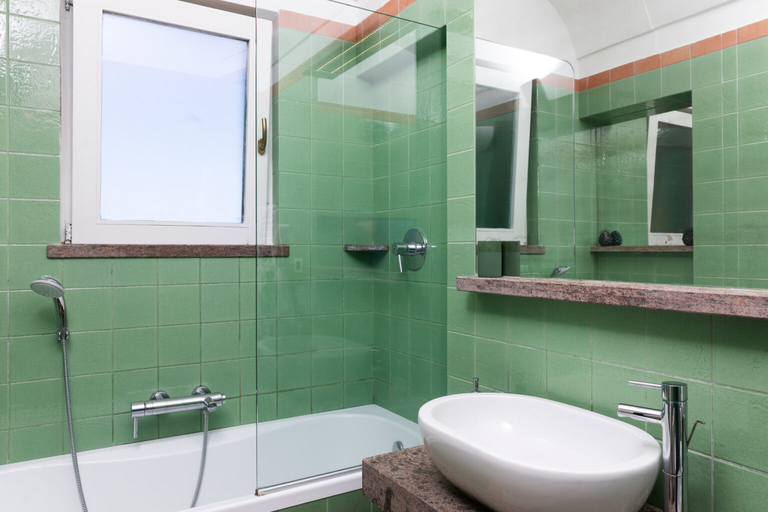 Modern bathroom featuring green tiled walls, a glass shower enclosure, a sleek white sink, and a bathtub. Natural light enters through a window, enhancing the contemporary design. | Only on SopranoVillas