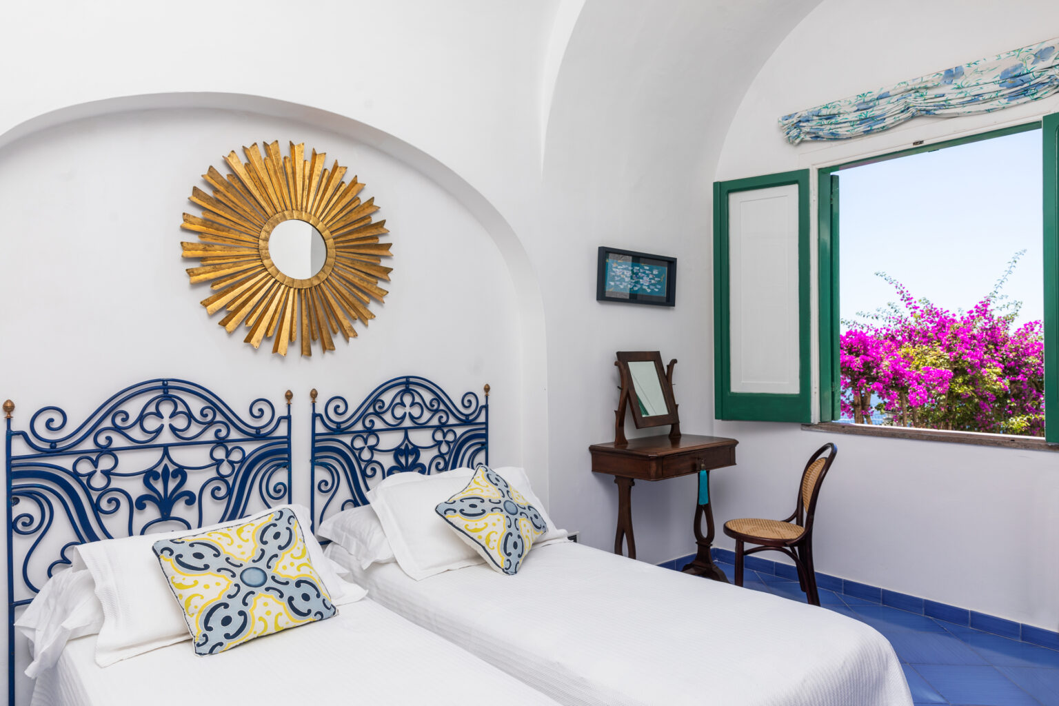 Bright and airy bedroom featuring two twin beds with decorative pillows, a vintage mirror on a wooden table, and a large window showcasing vibrant bougainvillea outside, all set against white walls and blue accents. | Only on SopranoVillas