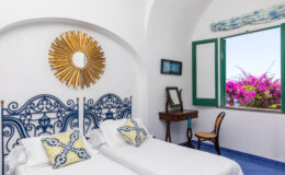 Bright and airy bedroom featuring two twin beds with decorative pillows, a vintage mirror on a wooden table, and a large window showcasing vibrant bougainvillea outside, all set against white walls and blue accents.