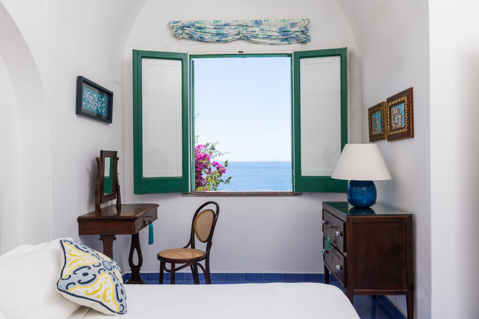 Cozy bedroom with white walls, green shutters, and a view of the ocean, featuring a wooden desk, a chair, and decorative accents. Bright natural light illuminates the space, highlighting the floral decor and inviting atmosphere. | Only on SopranoVillas