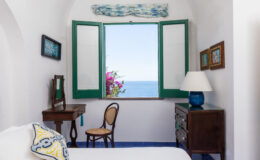 Cozy bedroom with white walls, green shutters, and a view of the ocean, featuring a wooden desk, a chair, and decorative accents. Bright natural light illuminates the space, highlighting the floral decor and inviting atmosphere.