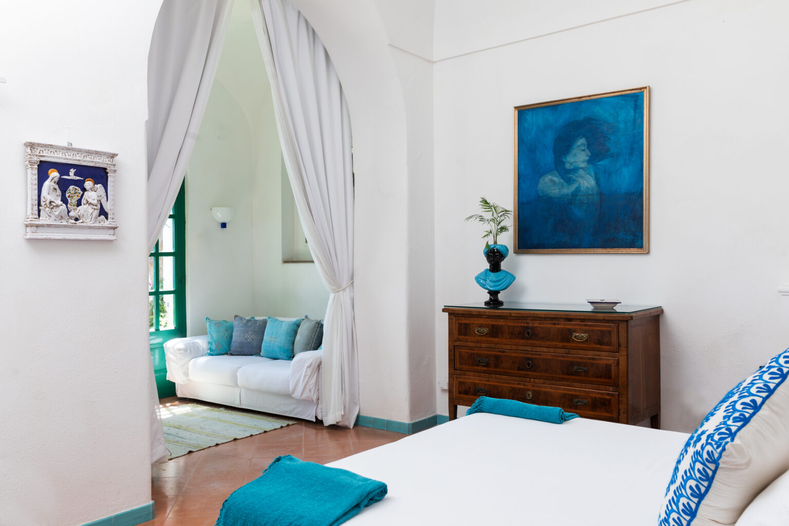 Bright and airy bedroom featuring a white bed with turquoise accents, a wooden dresser, and a cozy seating area in the background. Decorative artwork in blue tones adorns the wall, enhancing the room's serene atmosphere. | Only on SopranoVillas